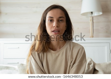 Millennial Caucasian girl sit on couch in living room look at camera having webcam conversation, young woman talk speak on video call using wireless internet connection at home, female blogger record Royalty-Free Stock Photo #1646348053