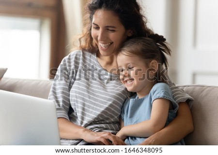 Head shot happy young curly mother embracing little preschool daughter, watching funny cartoons on computer in living room. Smiling mom enjoying free weekend time with cute child girl at home.