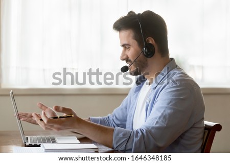 Millennial Caucasian male worker in headset talk consult client online using wireless internet on laptop, man call center agent or telemarketer in earphones speak with customer working on computer