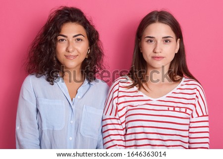 Horizontal shot of two females with calm face. Women standing isolated over pink stidio background, one lady wearing blue blouse, another striped shirt, looking directly at camera. People concept.