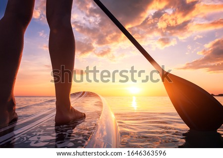 Stand up paddle boarding or standup paddleboarding on quiet sea at sunset with beautiful colors during warm summer beach vacation holiday, active woman, close-up of water surface, legs and board Royalty-Free Stock Photo #1646363596