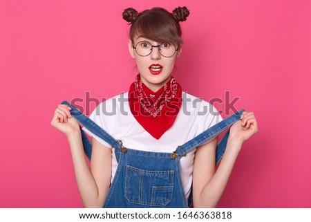Close up studio photo portrait of terrified astonished lady touching her overalls with hands, looking directly at camera, posing isolated over bright pink background. People emotions concept.