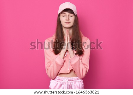Portrait of peaceful positive young female putting palms together, closing her eyes, having long brown hair, wearing pink hoodie, trousers and cap, praying alone, keeping silence. Peace concept.