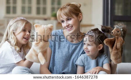 Happy young Caucasian mom play with little preschooler daughters use fluffy toys, smiling millennial mother or nanny engaged in doll theatre game with small girls children, have fun together at home