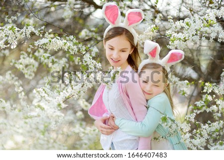 Two cute young sisters wearing bunny ears in blooming cherry garden on beautiful spring day. Kids hanging Easter eggs on blossoming cherry branches. Children celebratiing Easter outdoors.