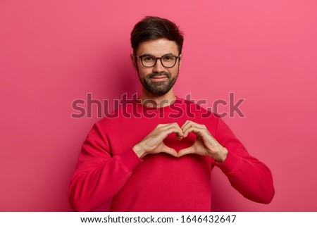 My heart belongs to you. Romantic handsome bearded man makes heart shape symbol with fingers, falls in love with woman, wears optical glasses and red sweater, isolated on pink studio background