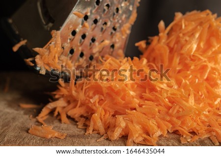 Grated carrots on a grater on the table. Closeup of a bunch of fresh uncooked root vegetables. Eye level shooting. Selective focus. Cooking process.