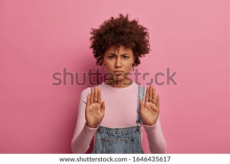 Portrait of serious strict woman does stop sign, has warning expression, shows limit and says enough, makes prohibition gesture, wears casual clothes, isolated on pink background, expresses denial