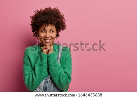 Headshot of dark skinned woman looks aside, has tender smile, keeps hands under chin, wears green sweater and dungarees, sees someting pleasant, isolated over rosy background. Emotions concept