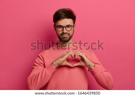 Handsome bearded man shares love with close people, shapes geart gesture, expresses sympathy and passion, heartwarming emotions, wears pink sweater, poses indoor. I love you with all my heart