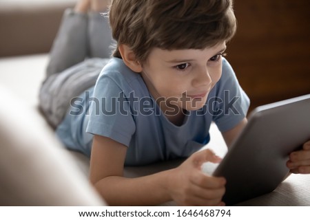 Focus on interested smiling little school boy playing online game on computer tablet. Happy small kid child using educational applications on electronic device or watching cartoons alone at home.