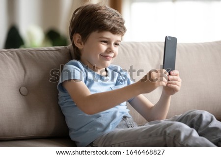 Funny little boy sitting on couch, taking selfie on smartphone. Happy small child resting alone on sofa, recording comedy video, watching cartoons, using mobile applications, web surfing alone at home