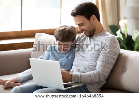 Overjoyed little school boy resting on sofa with smiling dad, enjoying funny movie on computer together at home. Excited happy two generations family laughing at cartoons, having fun indoors.