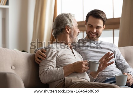 Happy middle aged grey haired man relaxing on cozy sofa with smiling grownup son, spending leisure weekend morning time with cup of coffee tea. Joyful two generations family talking sharing news. Royalty-Free Stock Photo #1646468869