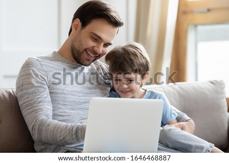 Joyful young father embracing little schoolboy son, showing applications on computer at home. Happy two generations male family enjoying funny movies cartoons on laptop together in living room.