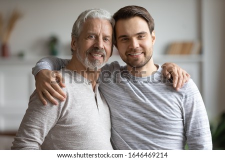 Head shot portrait pleasant elderly mature dad cuddling shoulders of grownup son. Affectionate happy two male generations family supporting each other, showing love care devotion, looking at camera.