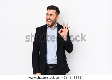 Young caucasian business man against a white background isolated winks an eye and holds an okay gesture with hand.