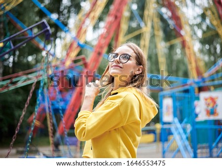 Young blond woman, wearing yellow hoody and blue jeans,spending time in amusement theme park in summer. Close-up portrait of pretty girl,standing near colorful ferris wheel, smiling. Leisure time