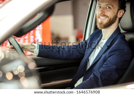 smiling caucasian guy riding new automobile which he bought in dealership, guy in formal suit enjoy sitting in new salon of car, sit behind the wheel. side view