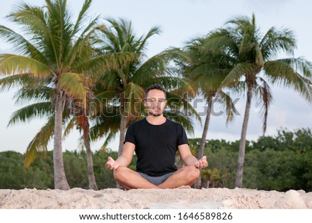 Young yoga man wearing black t-shirt at the beach, relaxing, breathing and meditating in a natural environment with palm trees and ocean as background. 