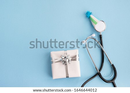 Doctor's day and World health day. Medical stethoscope in a festive cap and gift on light blue background. Creative medical postcard. Top view, flat lay, copy space
