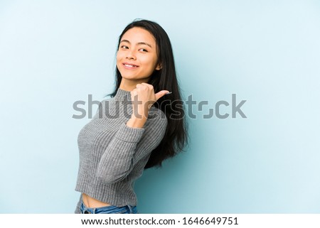 Young chinese woman isolated on a blue background points with thumb finger away, laughing and carefree.