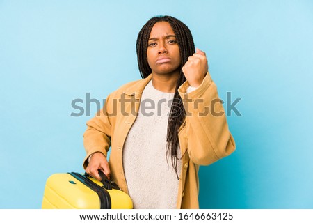 Young african american traveler woman holding a suitcase isolated showing fist to camera, aggressive facial expression.