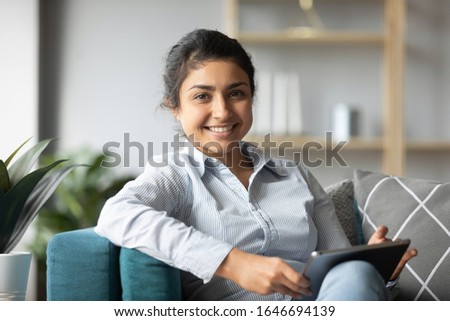 Profile picture of smiling millennial Indian girl sit on couch at home using modern tablet, portrait of happy young ethnic woman rest on sofa in living room shopping online browsing Internet on pad