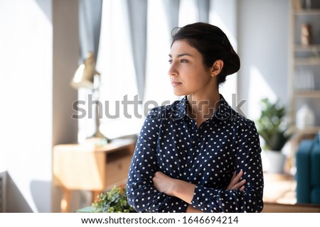 Pensive millennial Indian girl stand in living room look in window distance thinking or making decision, thoughtful young ethnic woman lost in thoughts pondering planning, remembering or recollecting Royalty-Free Stock Photo #1646694214
