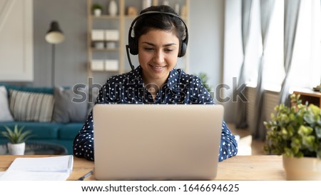 Smiling millennial Indian girl in wireless headset sit at desk in living room study online on laptop, happy young ethnic female in headphones watch webinar or course on Internet on computer Royalty-Free Stock Photo #1646694625