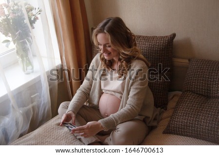 Beautiful  pregnant woman holding ultrasaund picture on her  tummy at home interiors. Close-up female hands keeping scan. Happy parent dresed beige home clothes and sit on bed. Maternity prenatal 