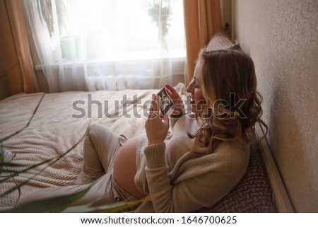 Beautiful  pregnant woman holding ultrasaund picture on her  tummy at home interiors. Close-up female hands keeping scan. Happy parent dresed beige home clothes and sit on bed. Maternity prenatal 