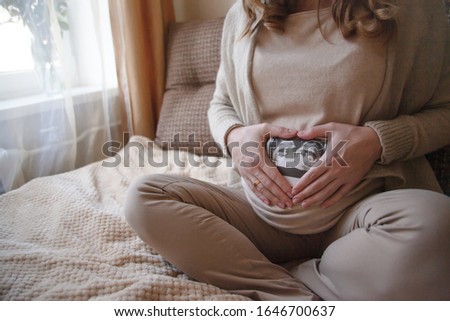 Cropped shot of pregnant woman holding ultrasaund picture on her  tummy at home interiors. Close-up female handsin heart keeping scan. Maternity prenatal 