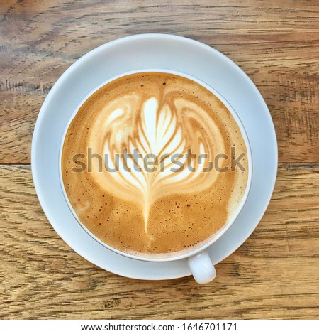 A white coffee cup of latte on a light brown wooden table, with latte art in the shape of a flower in the mousse.