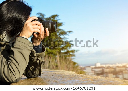 Young caucasian woman photographer taking picture of the city leaning on a wall on the top of a hill.