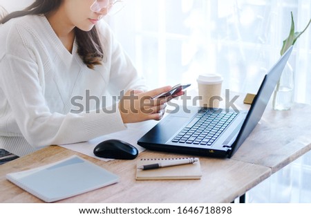 Close up of hand young female using mobile phone to searching information while she working with laptop on desk