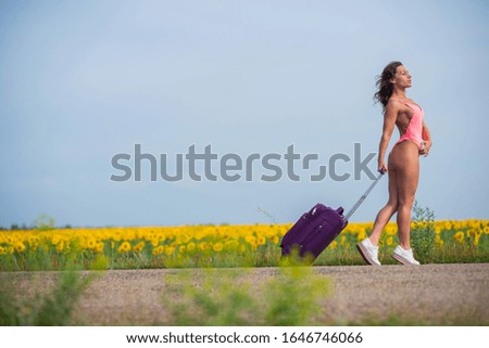 brunette in a pink monokini walks along a country road a field of sunflowers. Woman in a seductive swimsuit in the countryside with a luggage bag.