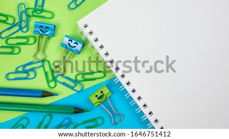 Office Paper Clip, Smile Binder Clips and Pencils on Notepad on Green and Blue Background. Open spiral notebook on table. Office supplies or education concept. Back to school