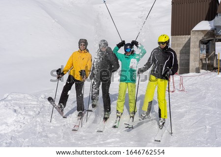 Young adults wearing skis, helmet and goggles, stand in line having fun, and holding sticks in the air on a sunny day in wintertime.