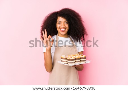 Young afro pastry maker woman holding a cupcakes isolatedYoung afro baker woman cheerful and confident showing ok gesture.