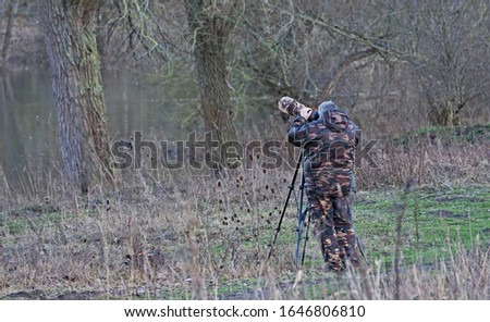 A man in camouflage suit is taking pictures with a camera on a tripod and a telelens covered in camouflage colours. Picture is low depth of field