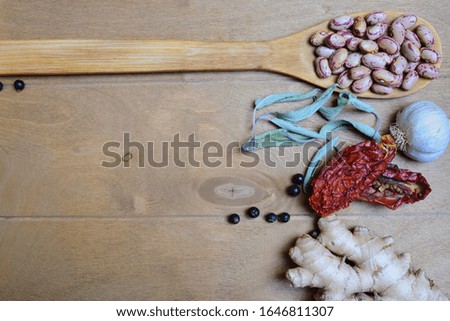 Top view of a wooden table with various ingredients for cooking, such as a spoonful of beans, juniper berries, sun-dried tomatoes, ginger, garlic and sage leaves