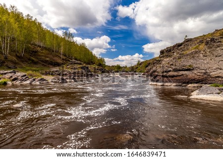 Summer landscape with fast-flowing river, stone banks, trees and blue sky with white clouds