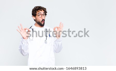 young doctor man feeling shocked, amazed and surprised, showing approval making okay sign with both hands against copy space wall