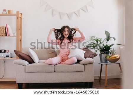Brunette with beautiful eyes smiles brightly and plays with her hair in bright, cozy apartment. Home portrait of girl dressed in pajamas sitting on beige sofa