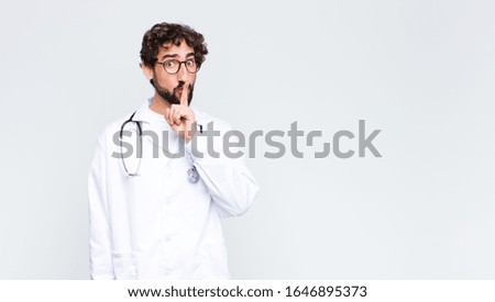 young doctor man looking serious and cross with finger pressed to lips demanding silence or quiet, keeping a secret against copy space wall
