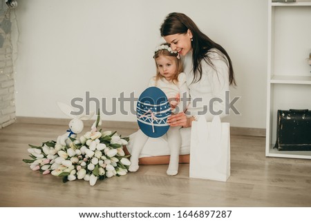 Mother with her daughter sitting on the floor looking at a white gift bag with a rabbit and a bouquet of flowers, and a painted egg, a Easter egg on white backround package 