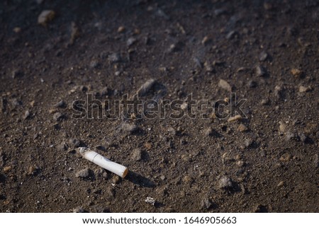 In the lower left corner of the picture is a smoking cigarette butt thrown on the asphalt. Asphalt background with small stones