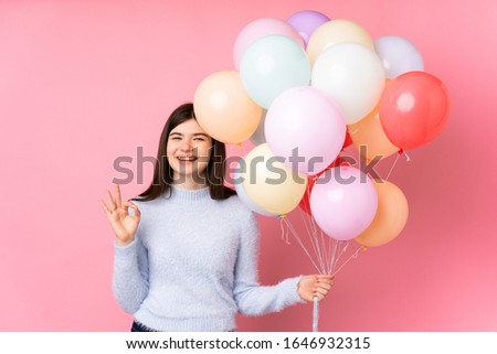 Young Ukrainian teenager girl holding lots of balloons over isolated pink background showing an ok sign with fingers