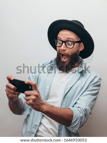 Handsome bearded guy holds smartphone and plays online games. Joyful man wearing a black stylish hat and a blue shirt. Crazy emotions.